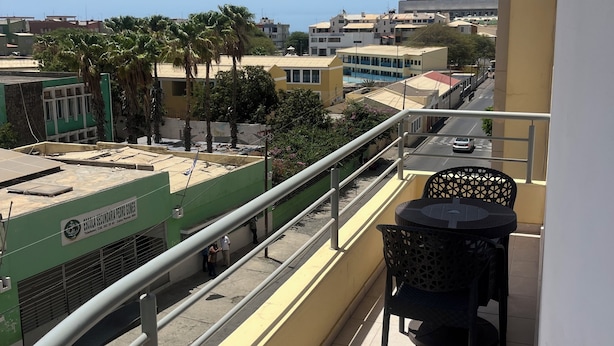 Un balcón con mesa y sillas, con vista a una calle con edificios y palmeras.