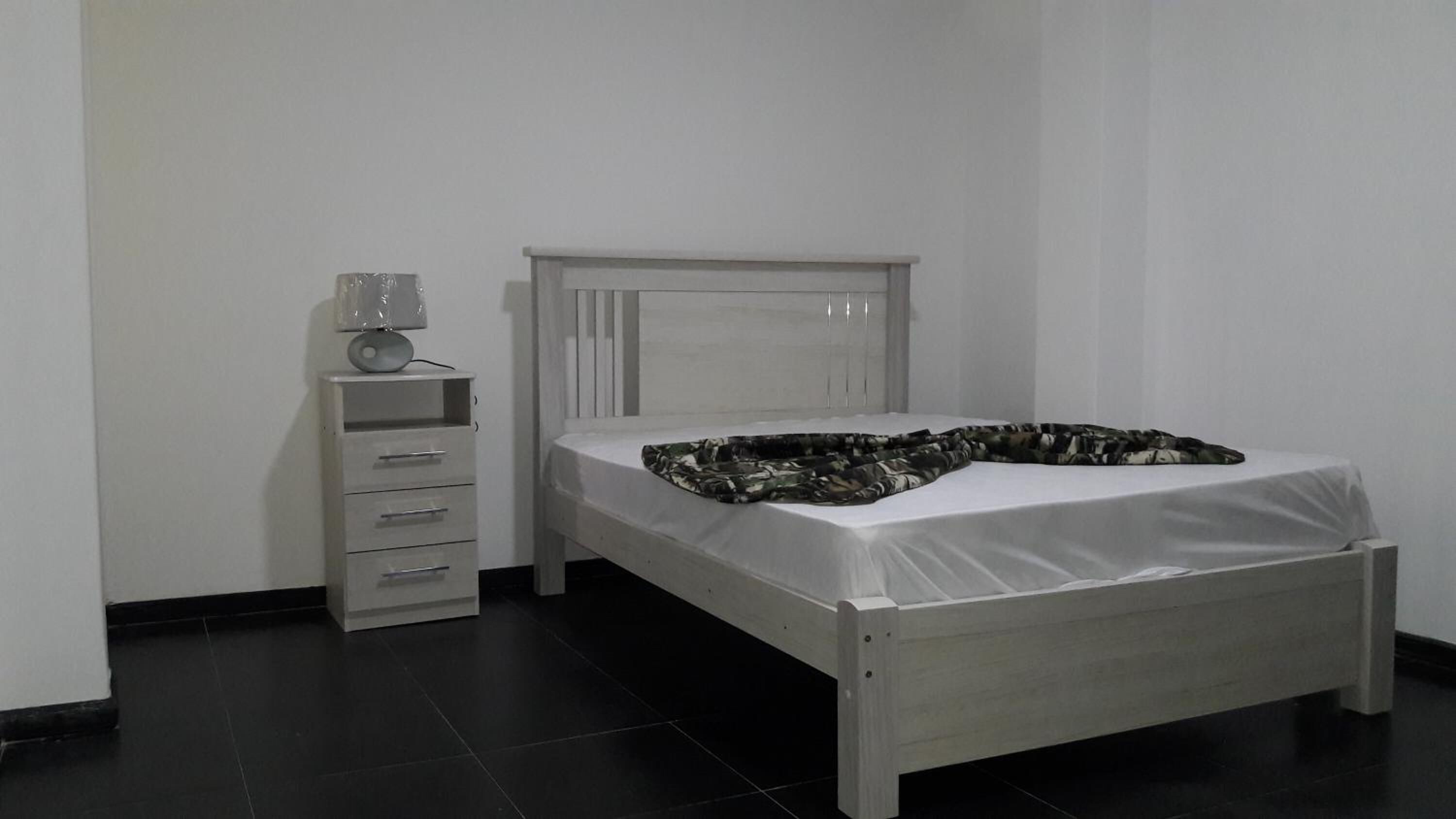 A single bed with a white mattress and a camouflage-patterned blanket, a white bedside table with drawers, and a white headboard.