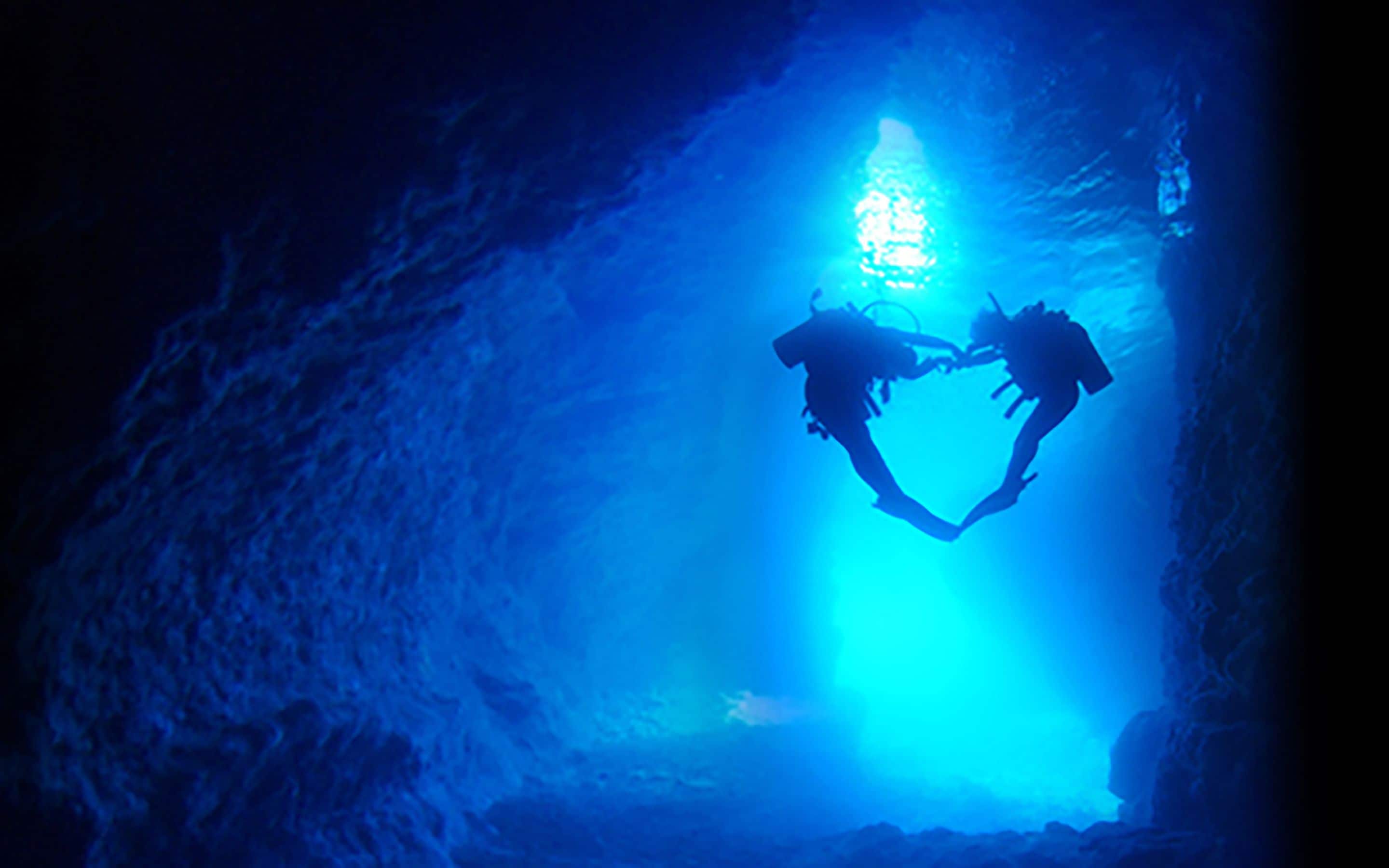 Twee duikers vormen een hart onder water in een grot, verlicht door blauw licht.