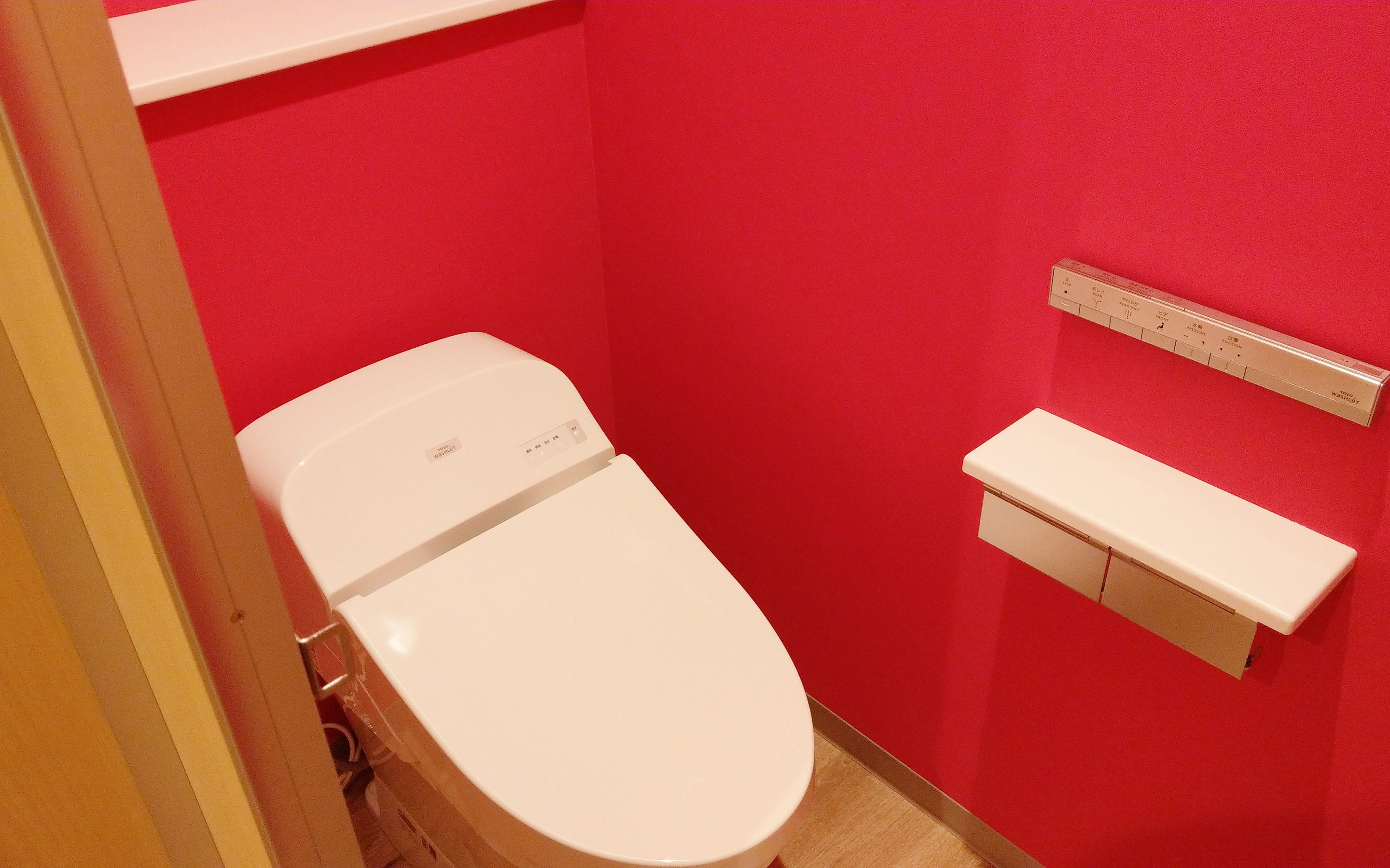 A small, red-walled bathroom with a white toilet, a white shelf, and a white toilet paper holder.