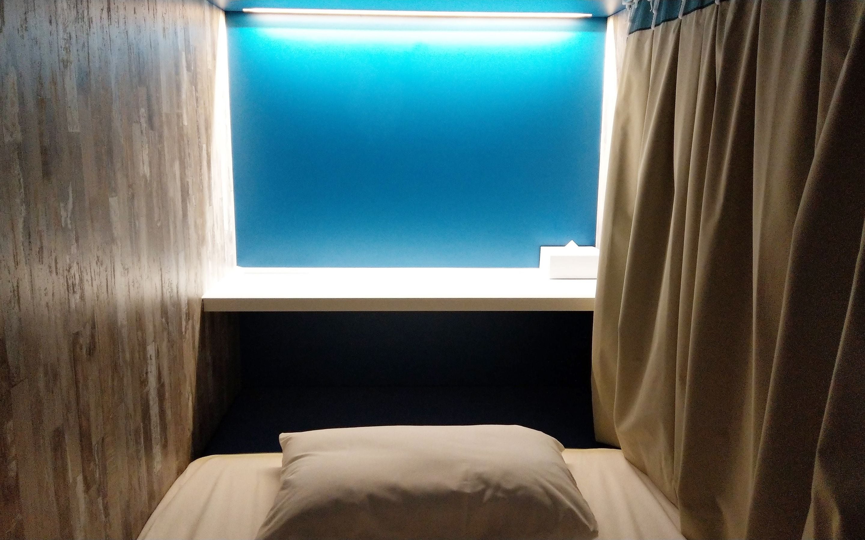 Una habitación pequeña y con poca luz, con una cama individual, un ventanal con tonalidad azul y una cortina.