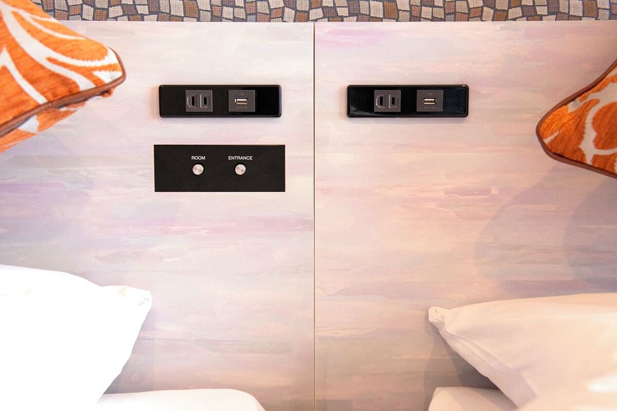 A hotel room with a bed, two USB charging ports, and a control panel with 'ROOM' and 'ENTRANCE' buttons.