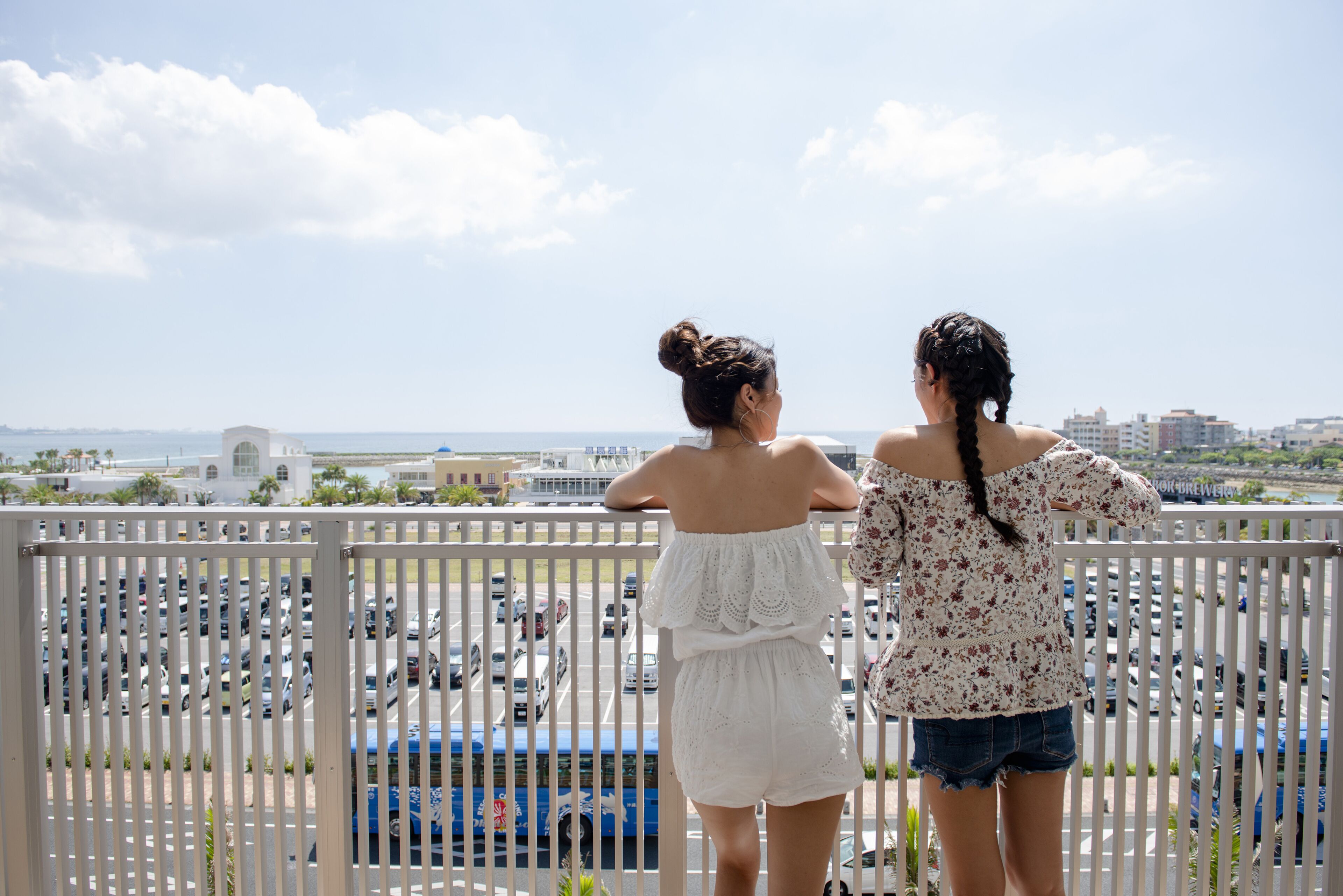 Zwei Frauen stehen auf einem Balkon mit Blick auf einen Parkplatz und eine entfernte Skyline.