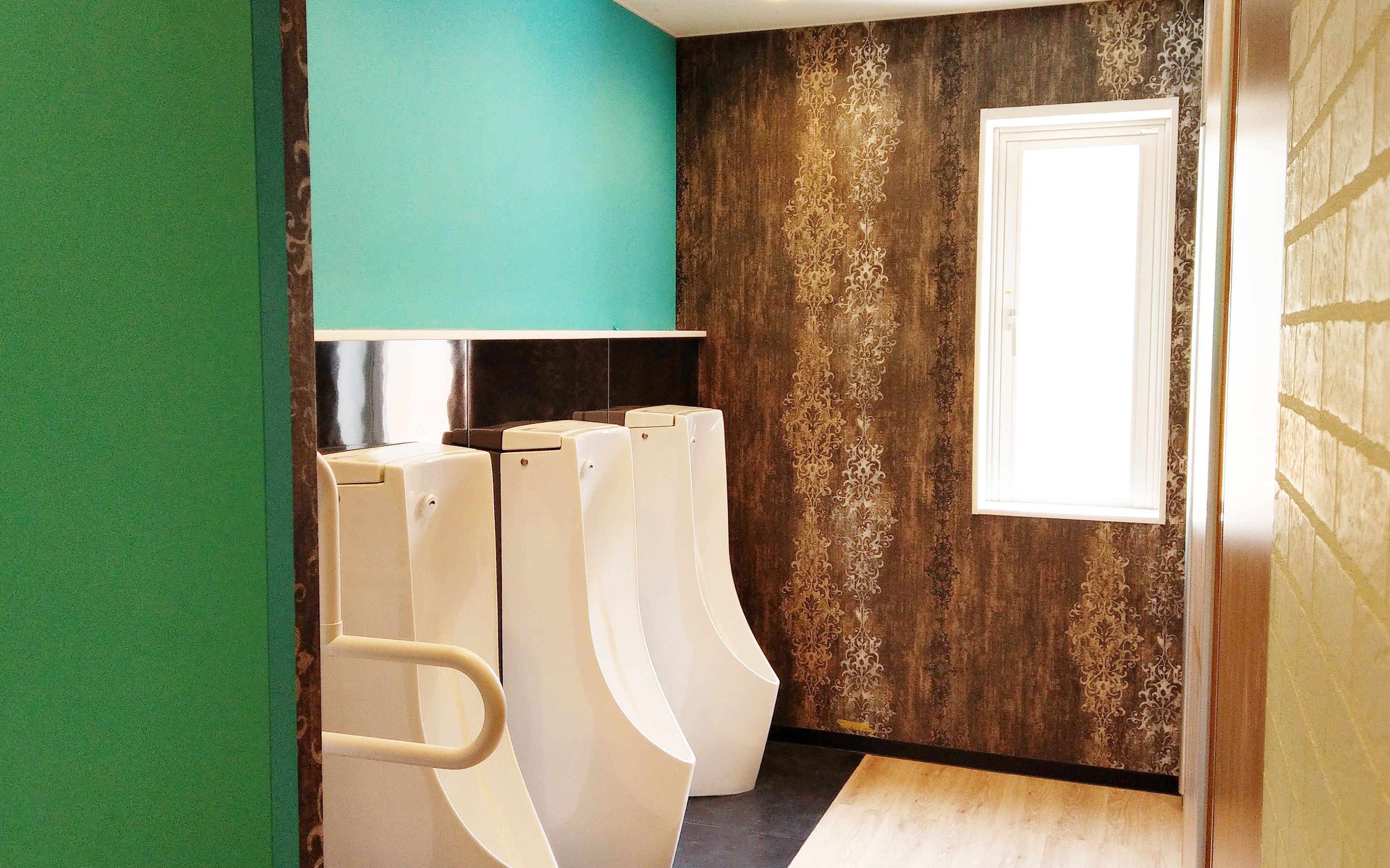 Un baño con tres orinales, una pared color verde azulado y una ventana con cortinas estampadas.