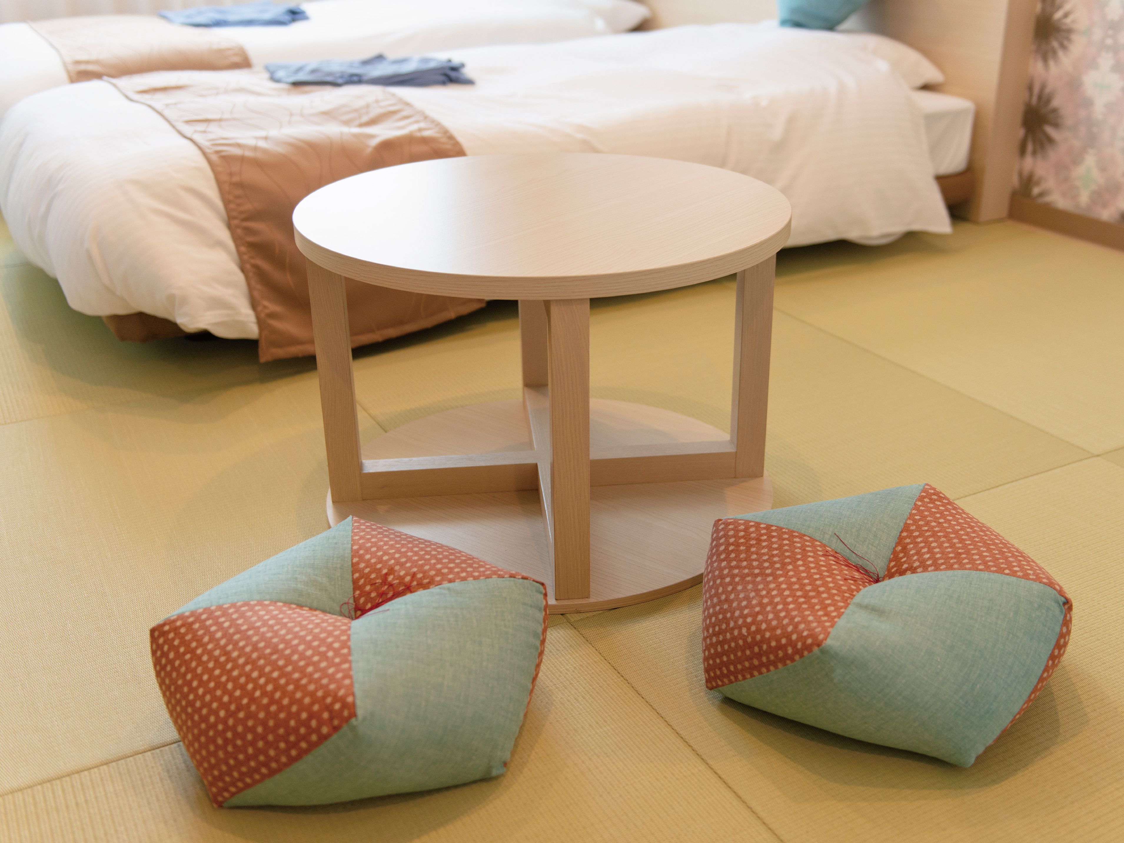 A room with a round wooden table, two matching cushions, and a bed with a white duvet and a brown blanket.