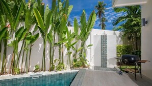 Uma área à beira da piscina com piscina azulejada, churrasqueira e plantas tropicais.