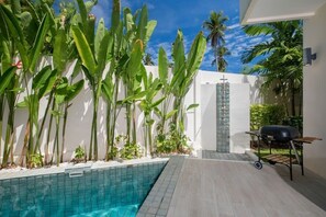 Un área junto a la piscina con piscina azulejada, una parrilla y plantas tropicales.
