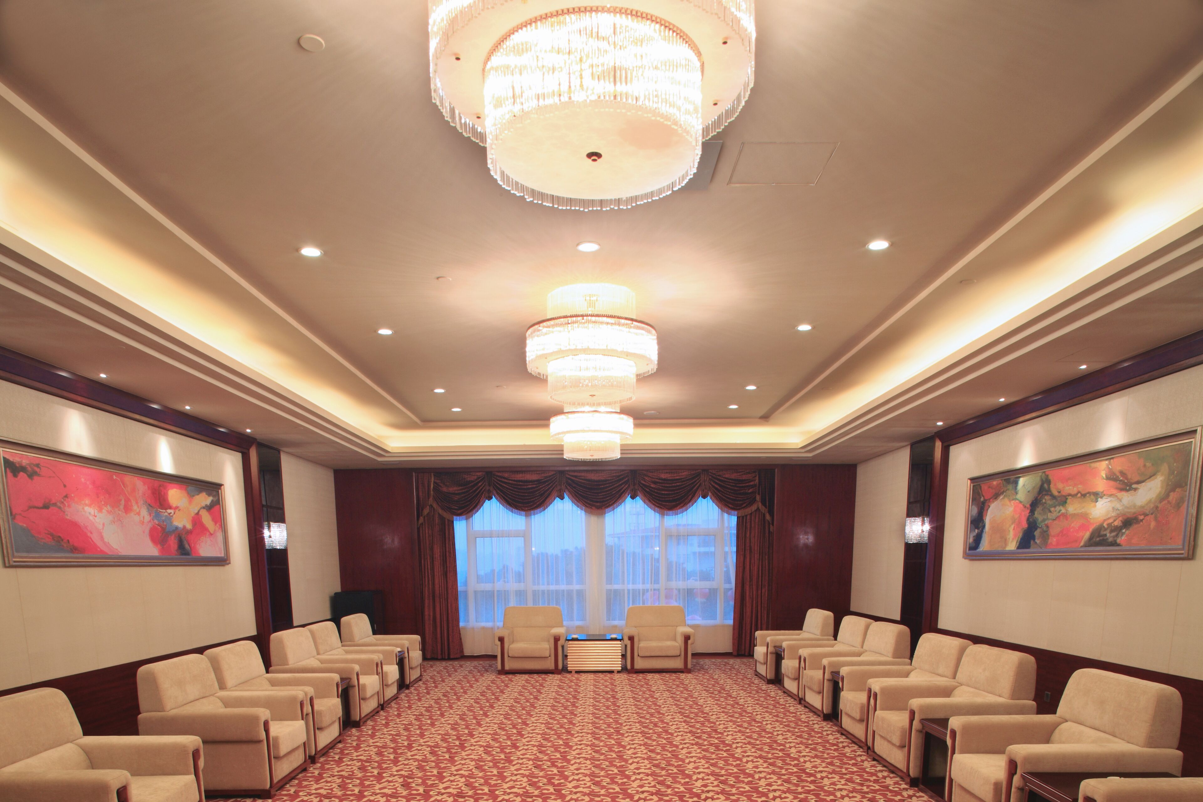 Un salón de conferencias con alfombra roja, varias filas de sillas color beige, una gran lámpara de araña y dos ventanas amplias con cortinas.