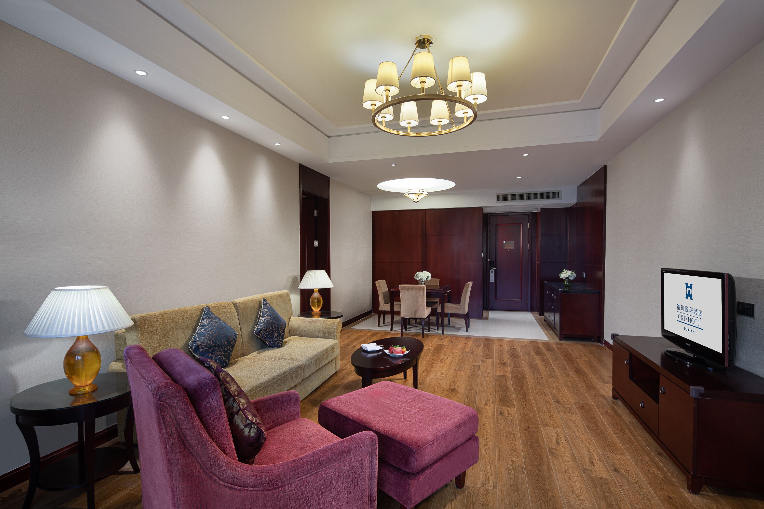 Quarto de hotel com sofá, poltrona, televisão, luminária, mesa de jantar e cadeiras.