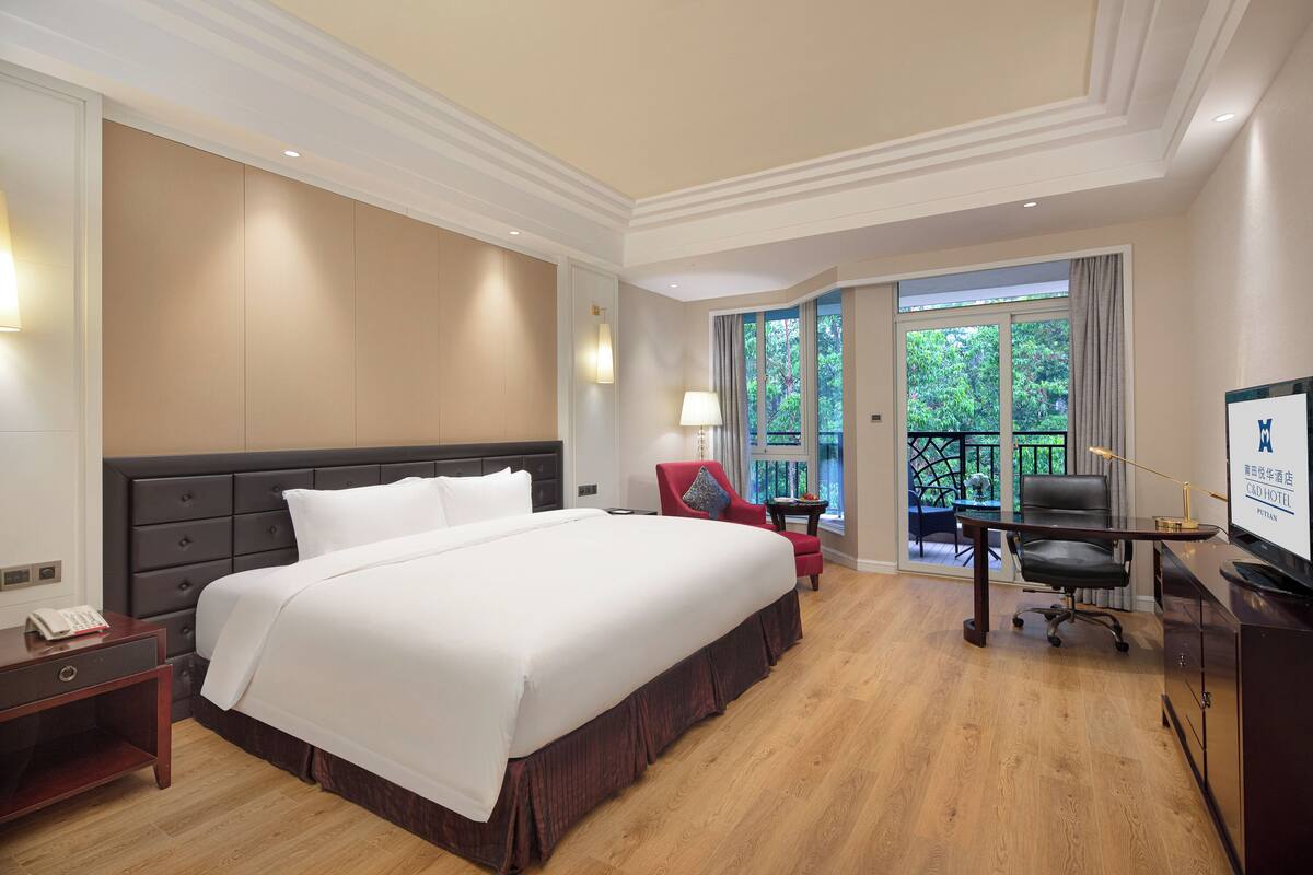 A hotel room with a large bed, a desk, a chair, and a balcony with a view of greenery.