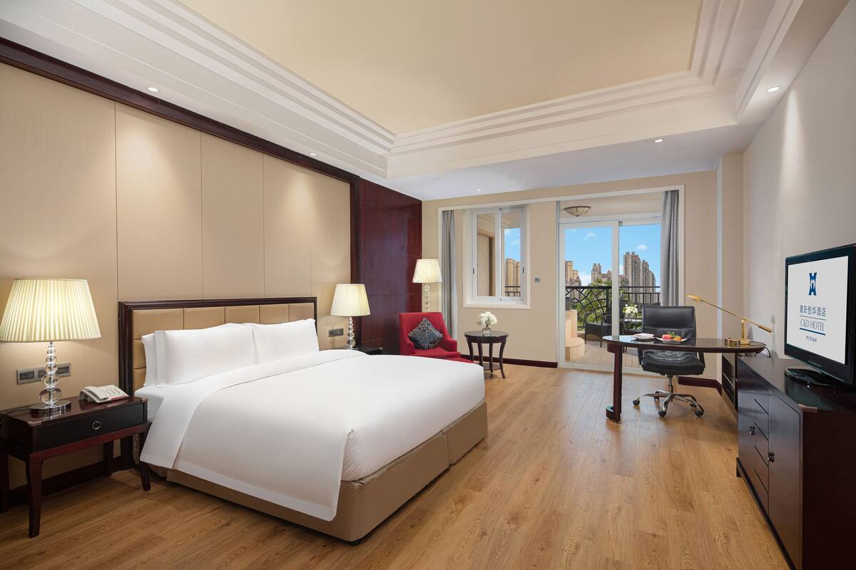 A hotel room with a large bed, a desk with a chair, a TV, and a balcony with a city view.
