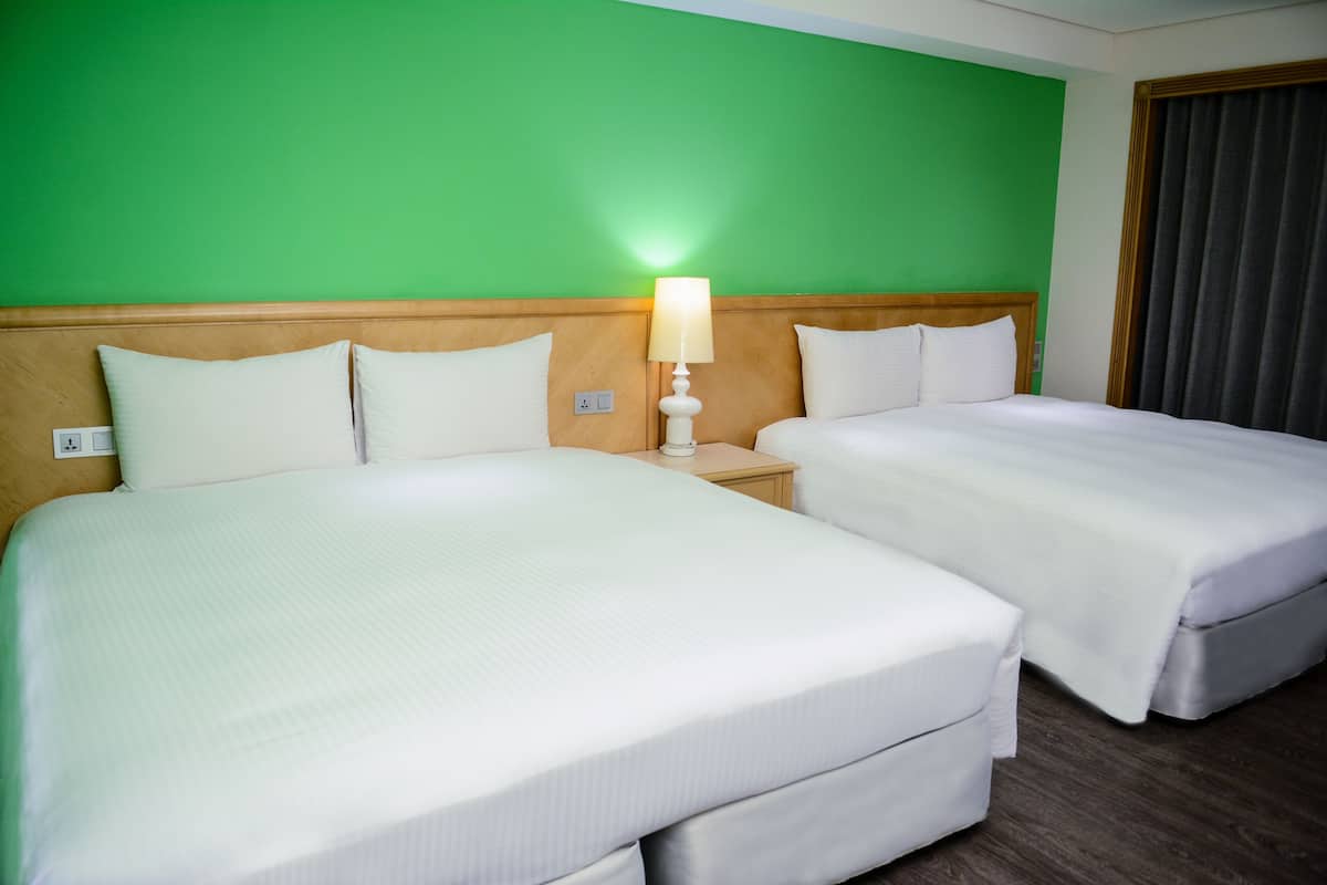 A hotel room with two beds, a green wall, wooden headboard, and a nightstand with a lamp.