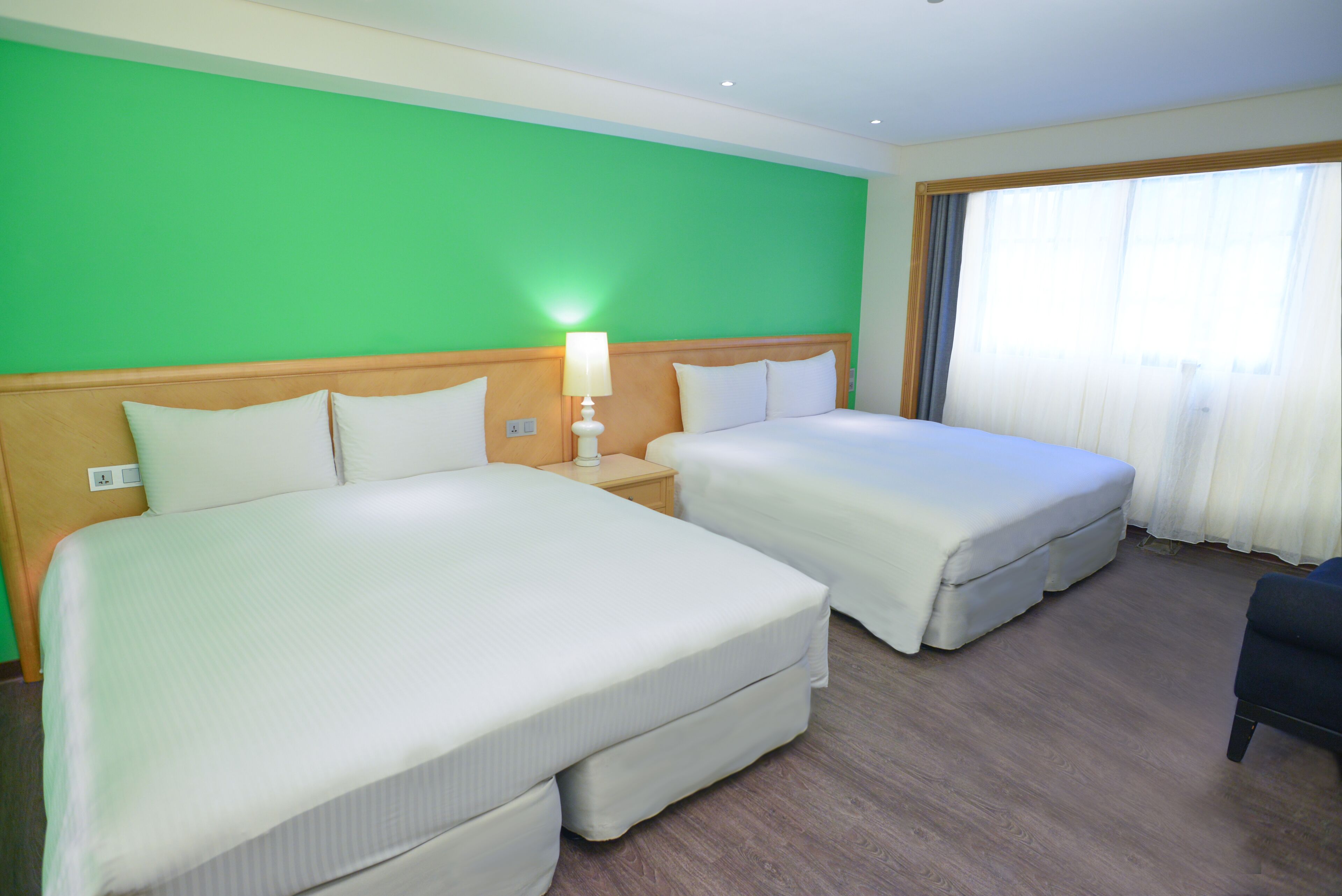 A hotel room with two beds, a nightstand with a lamp, and a large window with curtains.