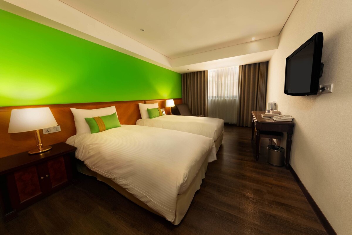 A hotel room with two beds, a television, a desk, and a trash can.