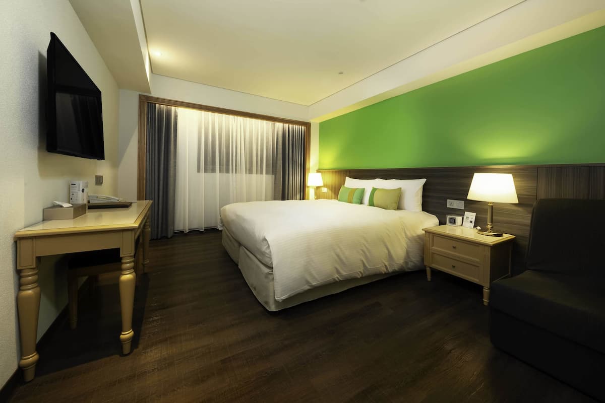 A hotel room with a bed, bedside tables, a desk, a chair, a TV, and a green accent wall.