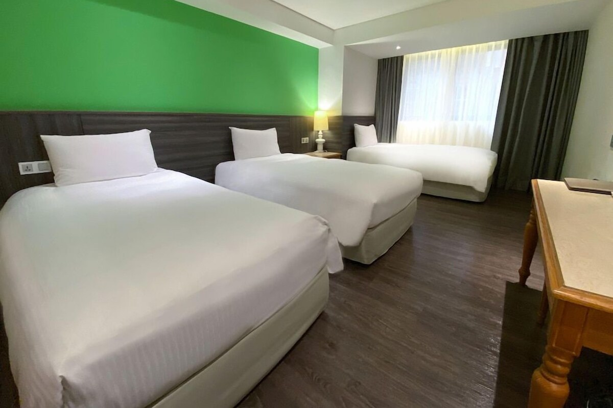 A hotel room with two beds, a desk, a green wall, and a window with curtains.