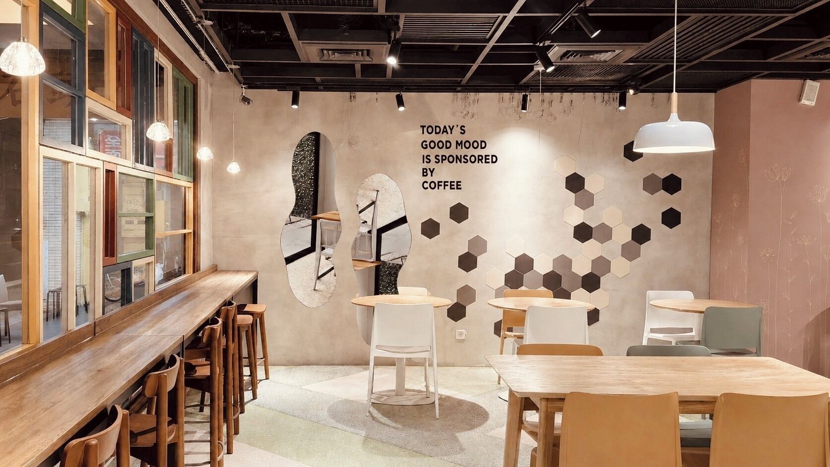 Interior de una cafetería moderna con mesas y sillas de madera, un reloj de pared grande y decoración mural hexagonal.