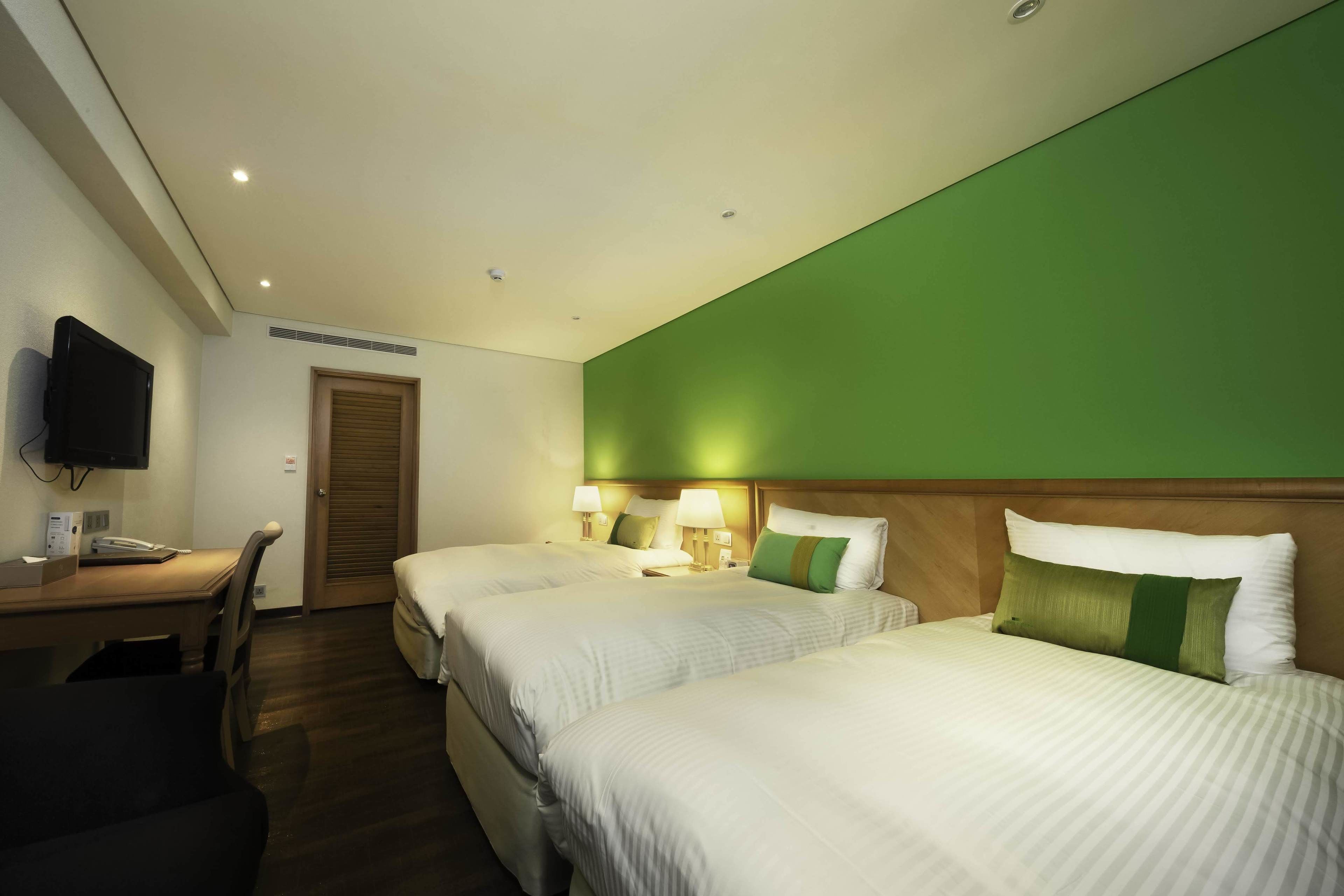 A hotel room with two beds, a desk, a television, and a green accent wall.