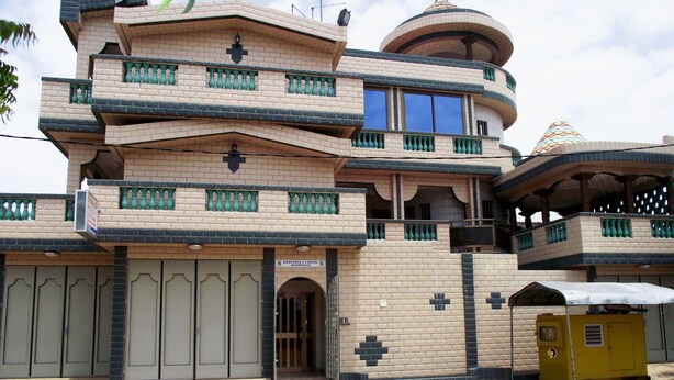 A multi-story hotel with a distinctive architectural design, featuring balconies with green railings and a central circular tower. - Hotel Magnificat Baguida (Lomé)