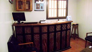 A reception desk with a computer monitor, a telephone, and a small table with a vase. There is a window with blinds and a door with a wooden frame. - Hotel Magnificat Baguida (Lomé)