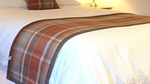 Ein ordentlich gemachtes Bett mit weißen Leinentüchern, einer Karo-Decke und einem einzelnen roten und braunen Kopfkissen.