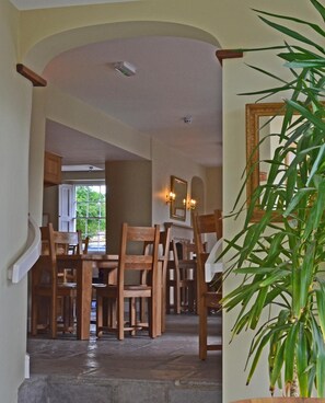 A dining area with wooden chairs and tables, a stone floor, and a view of a window with greenery outside. - The Drayton Crown (Langport)