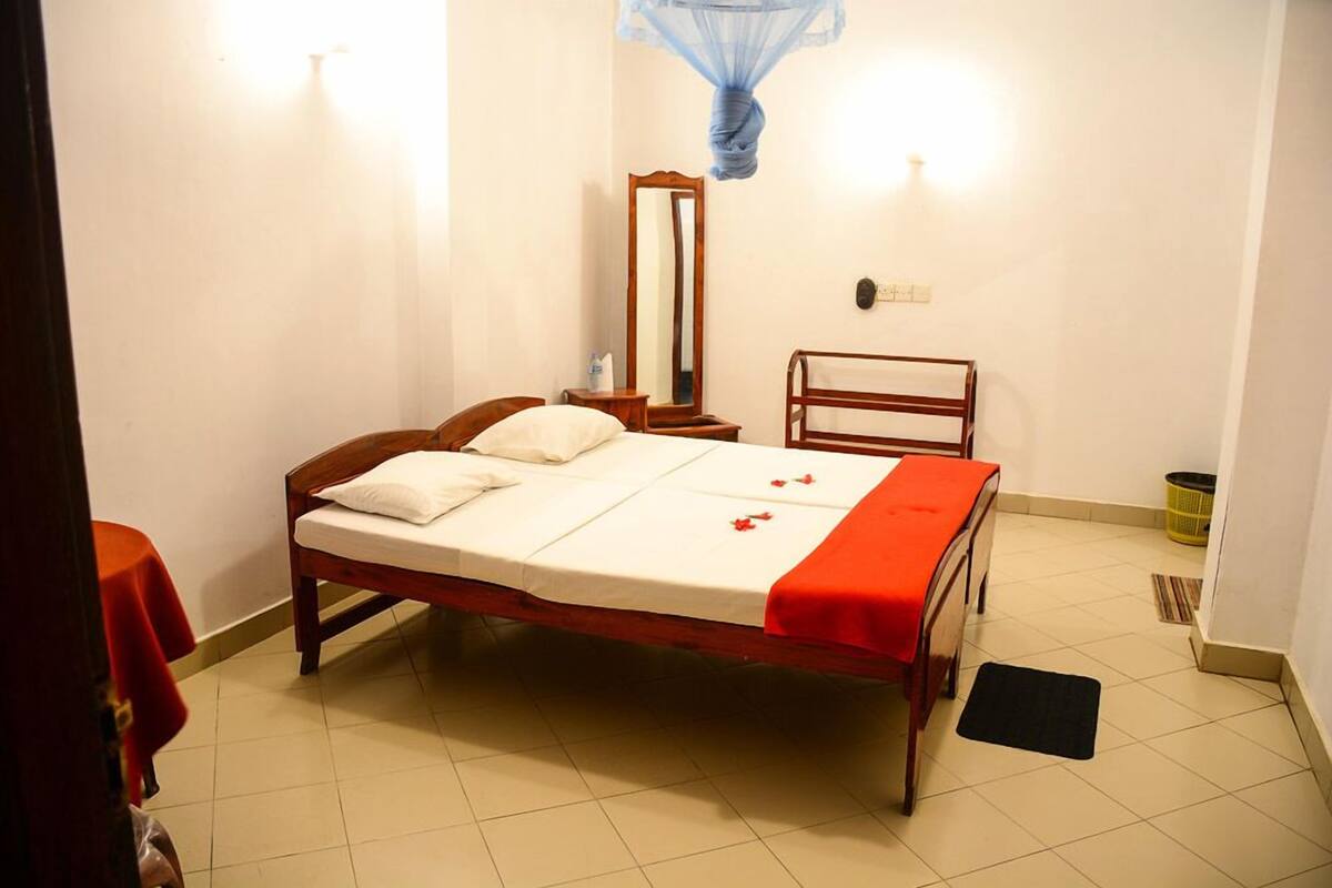 A neatly made bed with a white mattress and red bedding, a wooden headboard, a bedside table with a mirror, and a small red table beside the bed.