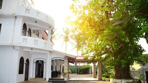 A white building with a covered patio area, a large tree, and a clear sky. - Shanthi beach resort (Unawatuna)