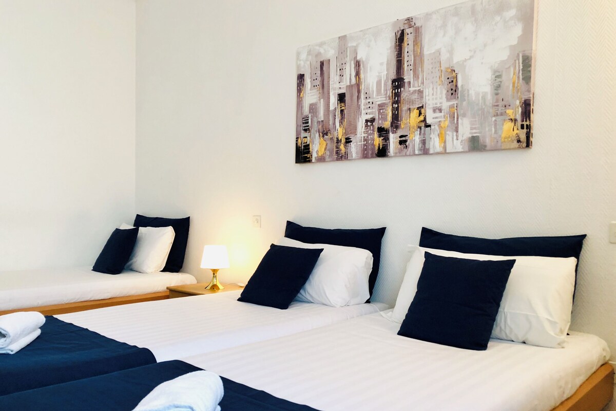A hotel room with two beds, white linens, dark blue bedding, and a cityscape painting on the wall.