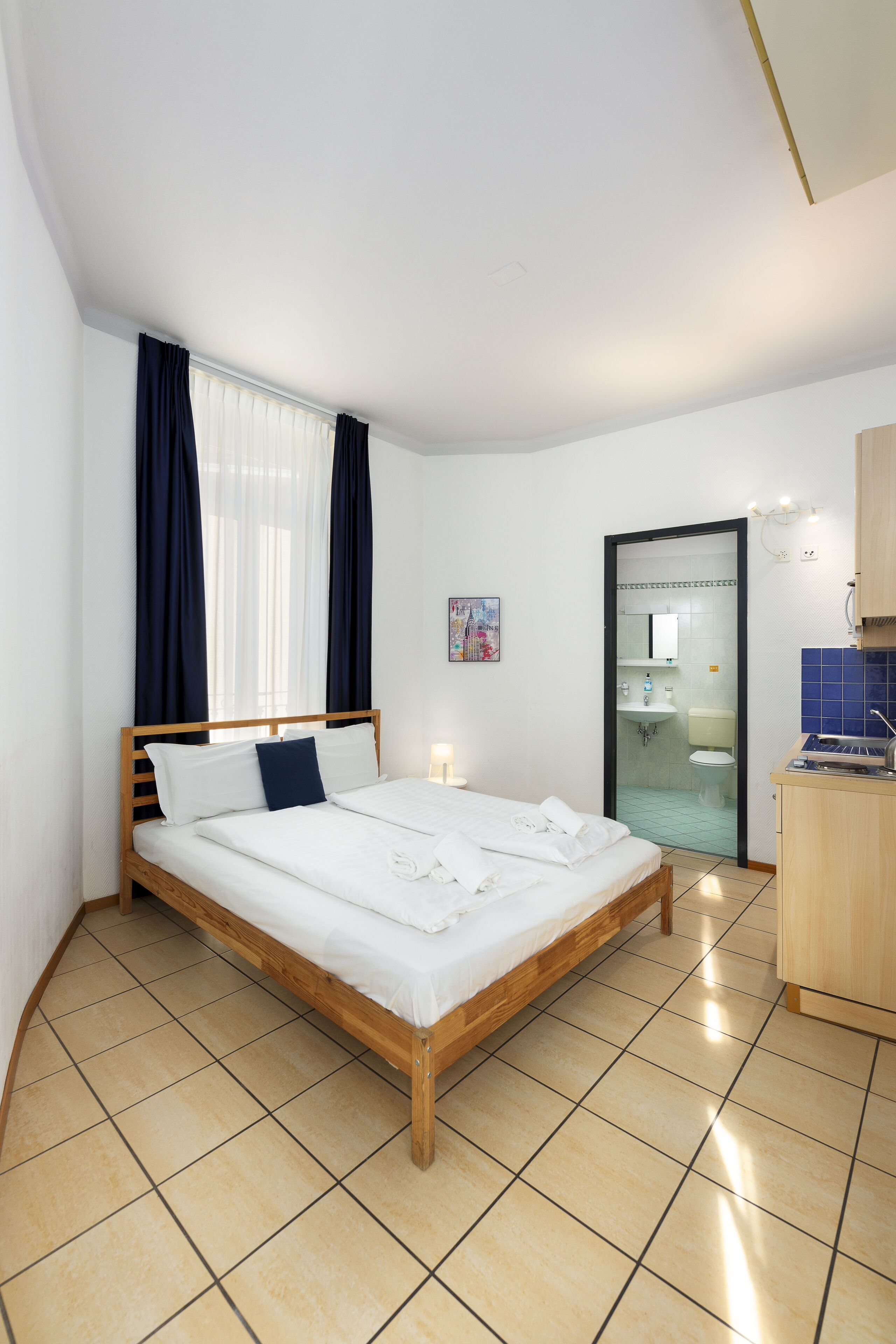 Une chambre d’hôtel moderne avec un lit en bois, une salle de bain visible à travers une porte en verre et une petite kitchenette.