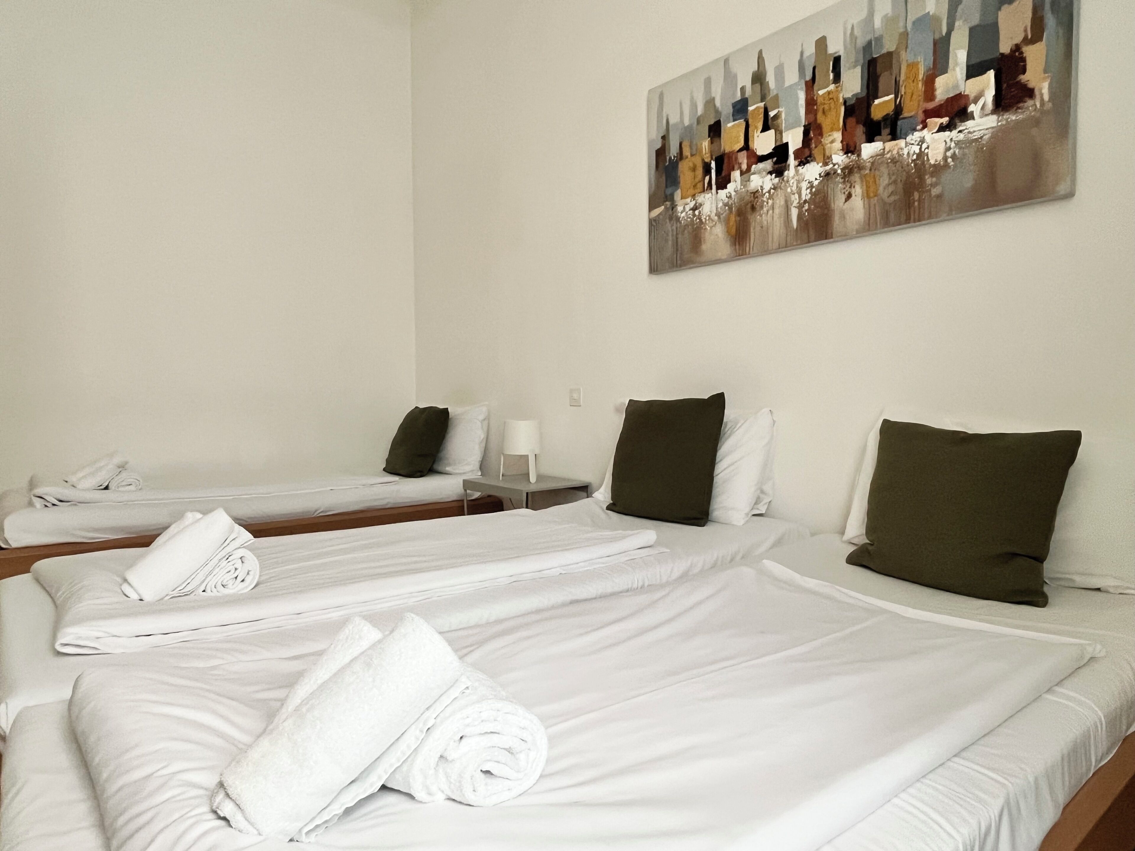 Une chambre d’hôtel avec trois lits, chacun doté de draps et d’oreillers blancs, un tableau au mur et une table de chevet avec une lampe.
