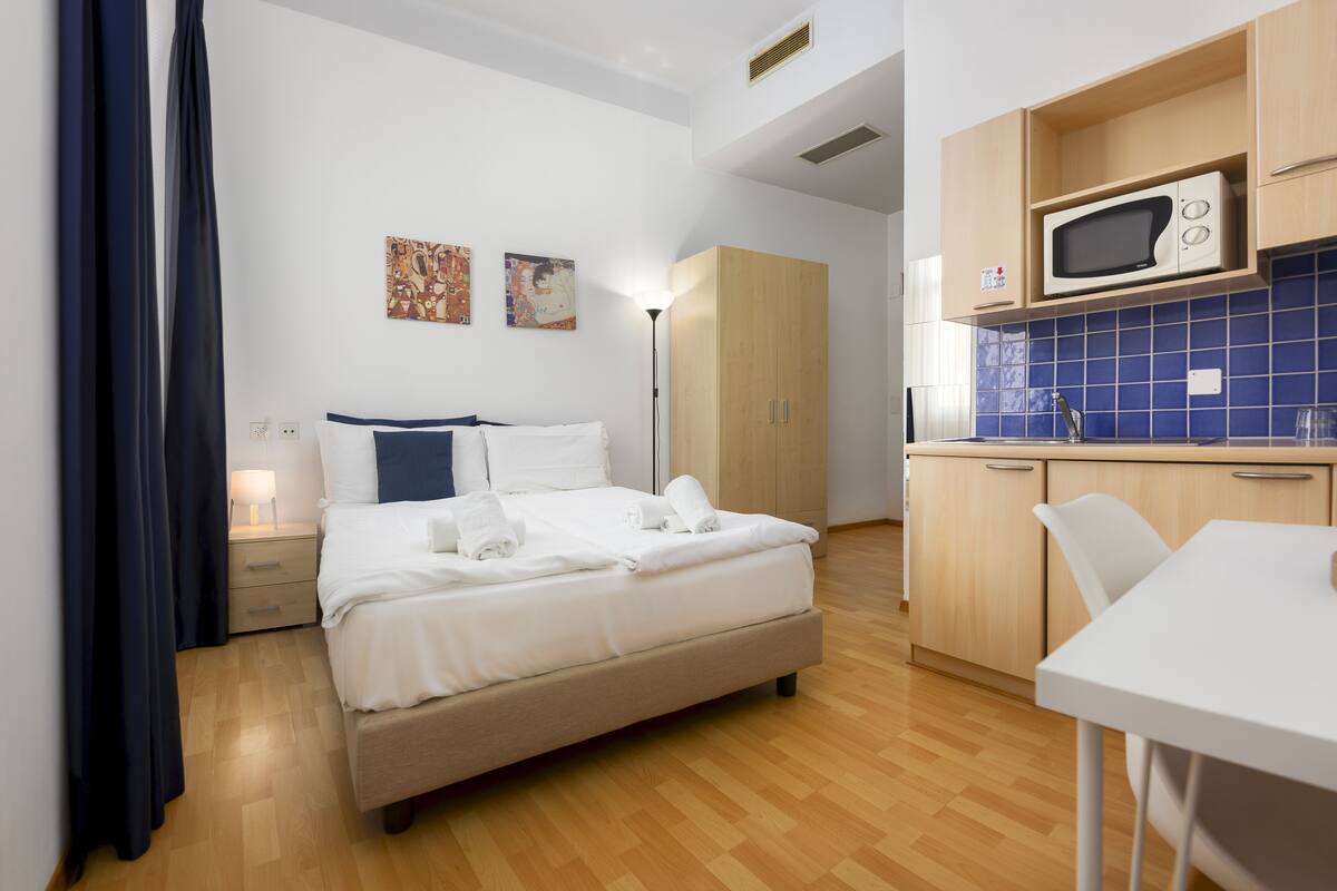 A hotel room with a bed, a bedside table, a lamp, a wardrobe, a kitchenette with a microwave, and a dining area.