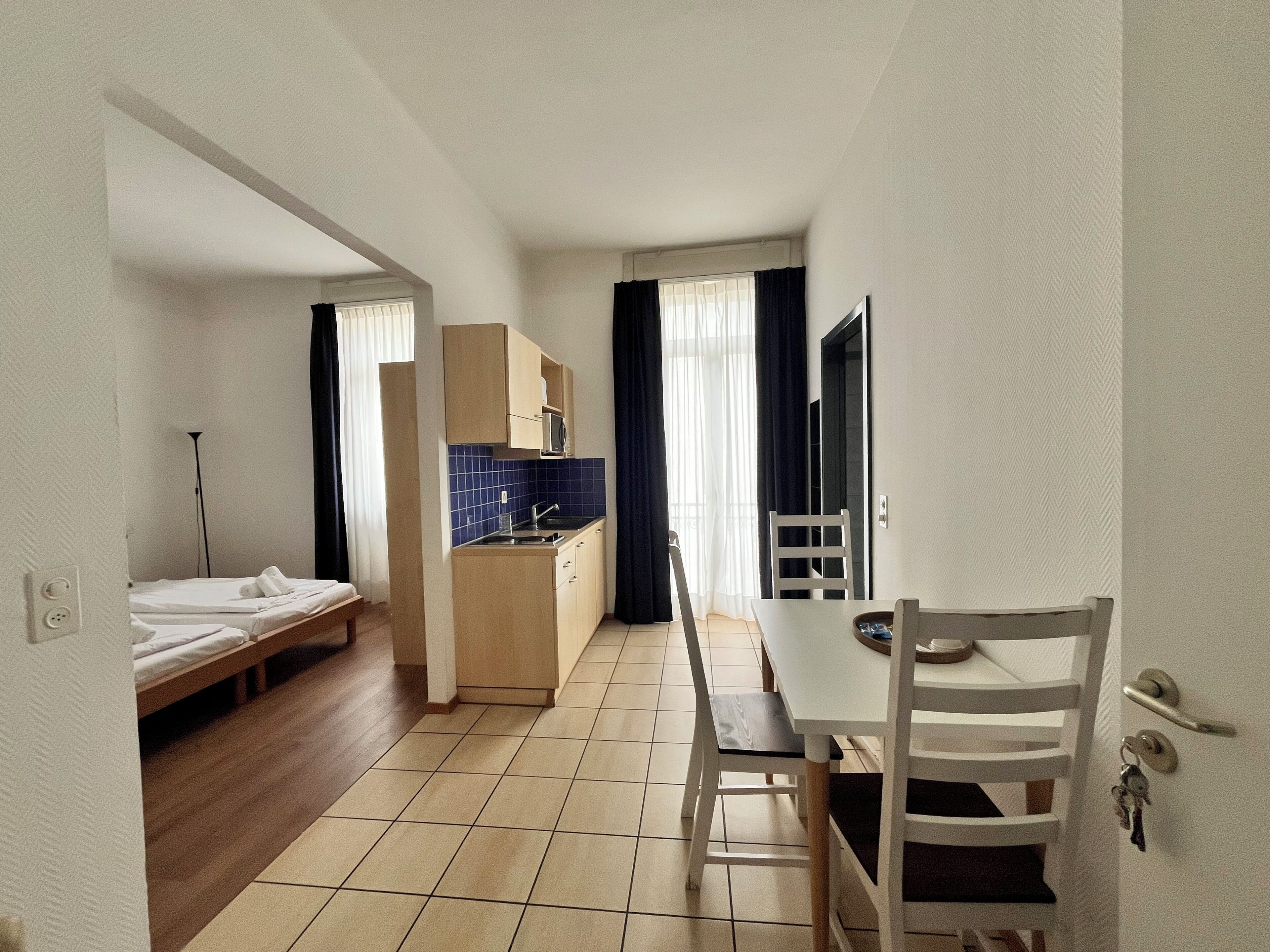 Une chambre d’hôtel compacte comprenant un coin cuisine, un lit simple, une table à manger avec des chaises et une petite fenêtre avec des rideaux.