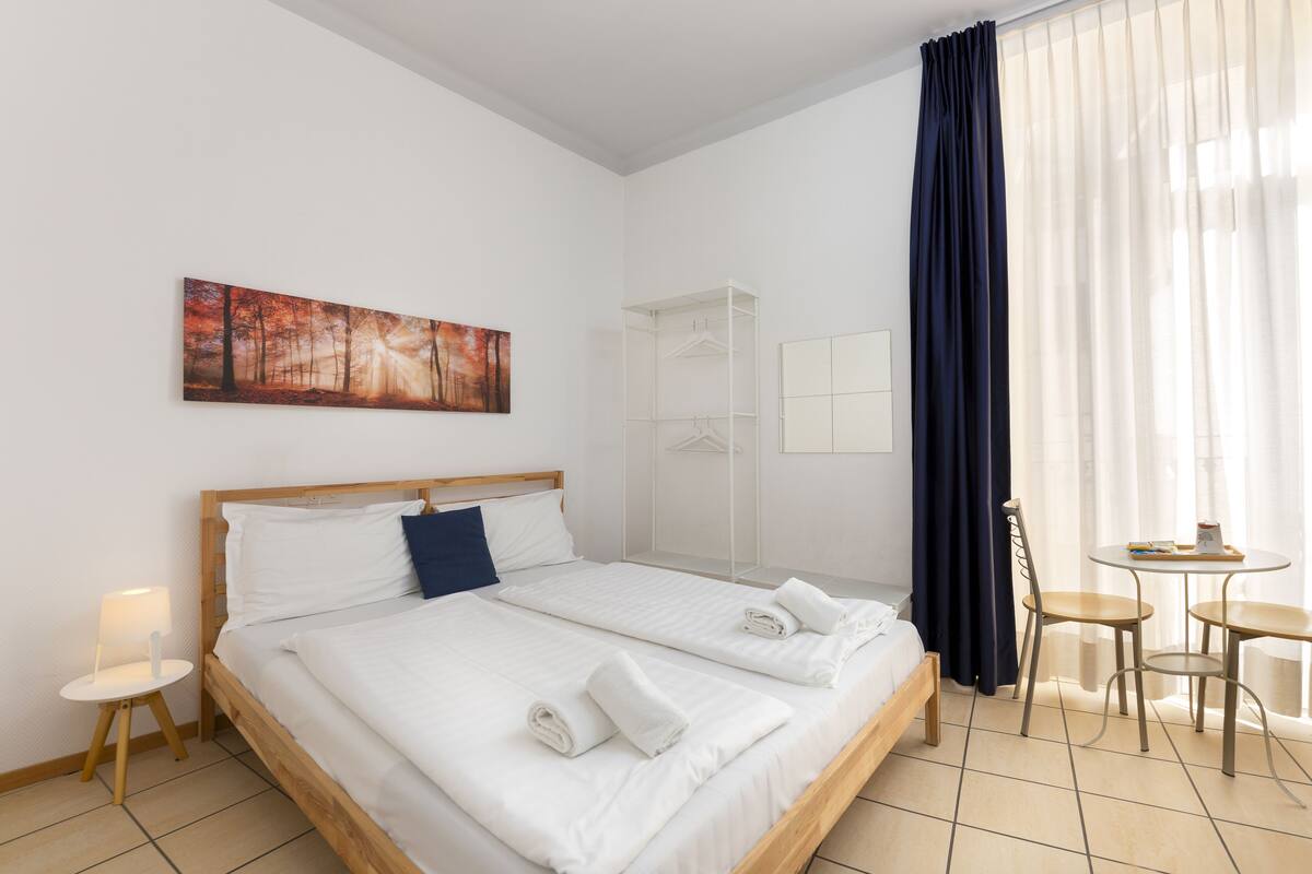 A hotel room with a large bed, a bedside table with a lamp, a chair, a small table, and a wardrobe.