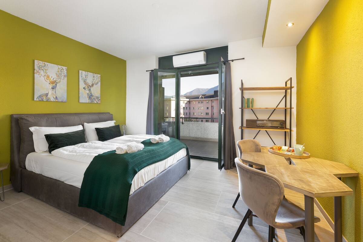 A hotel room with a bed, a dining table, a chair, and a balcony with a view of buildings.