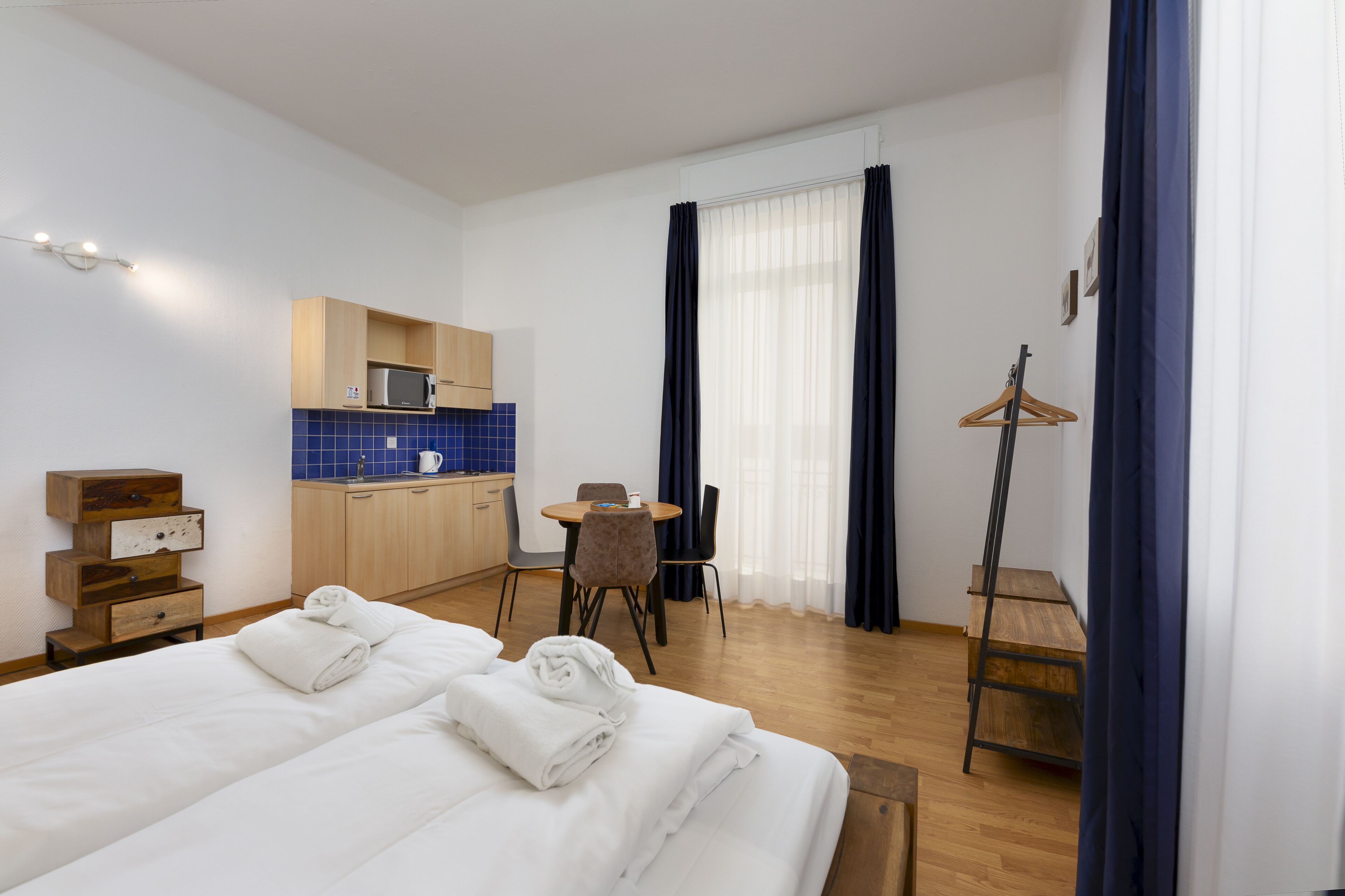 A hotel room with a bed, a desk, a chair, a small kitchenette, and a coat rack.