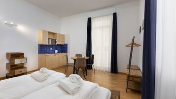 A hotel room with a bed, a desk, a chair, a small kitchenette, and a coat rack.