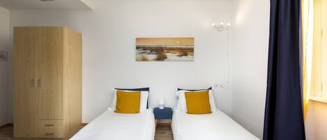 A hotel room with two beds, a wooden wardrobe, a painting on the wall, and a window with curtains.