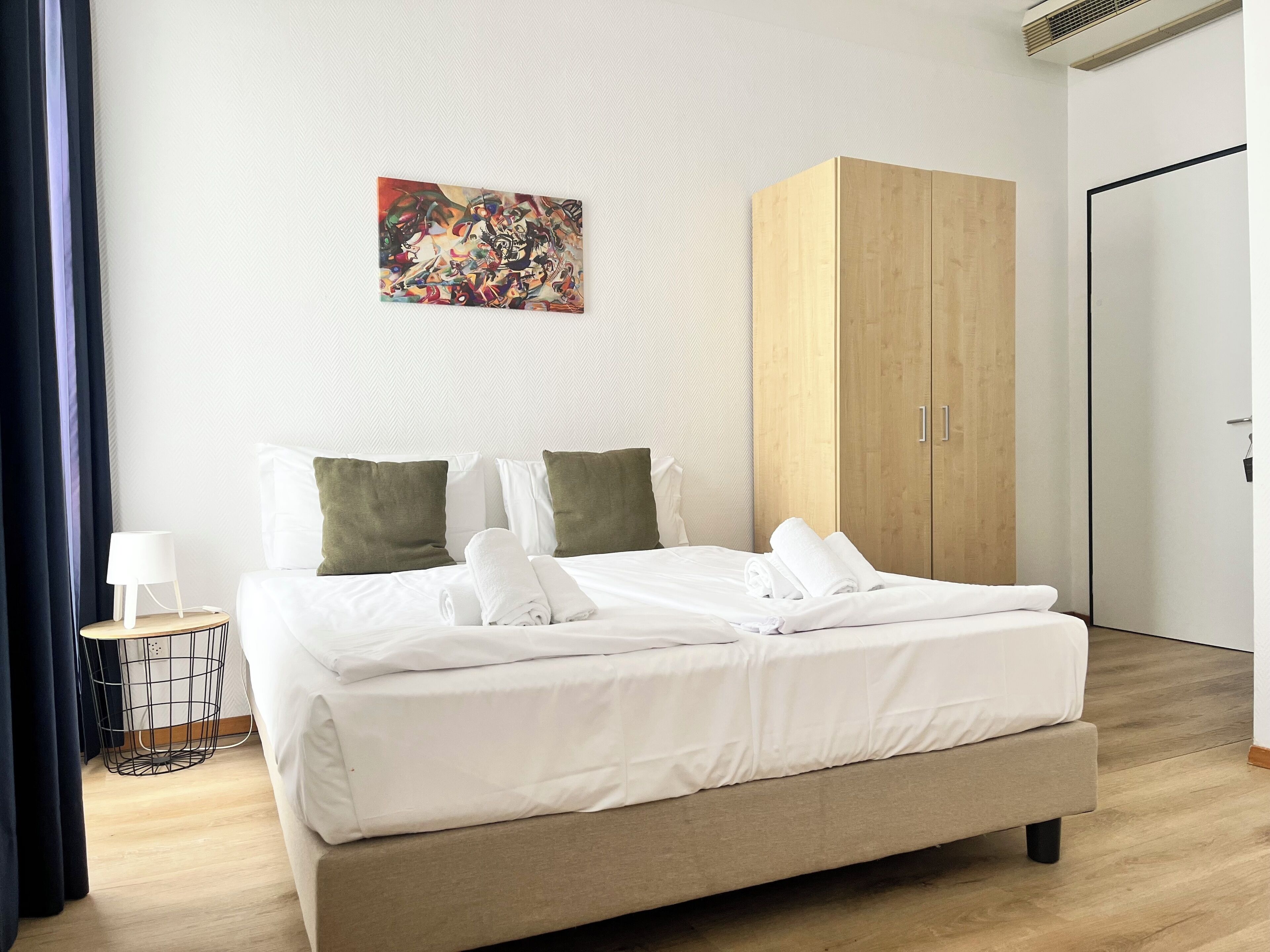 A neatly made bed with white linens, a wooden wardrobe, and a small bedside table with a lamp.