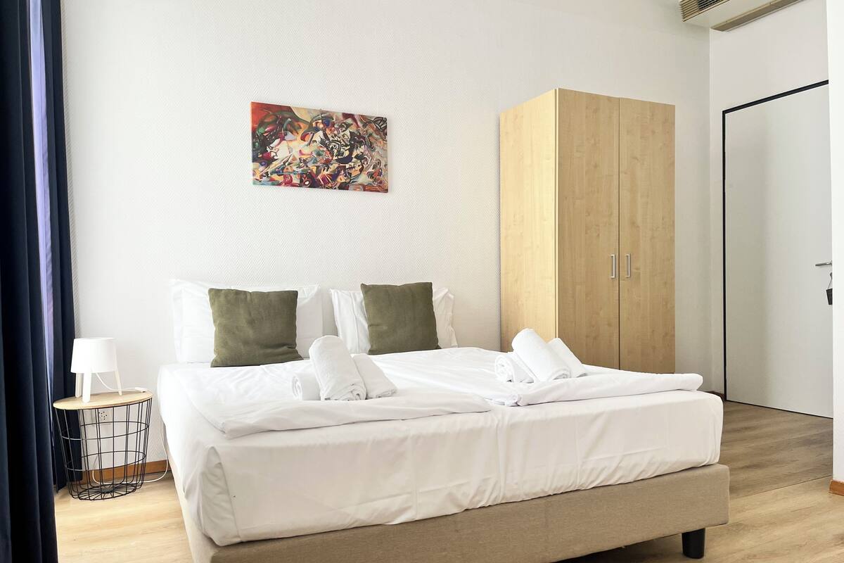 A neatly made bed with white linens, a wooden wardrobe, and a small bedside table with a lamp.