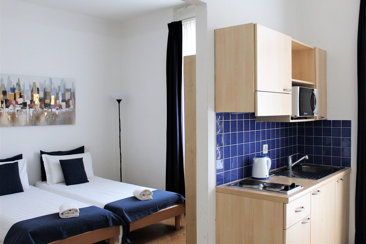 A hotel room with two beds, a small kitchenette, and a painting on the wall.