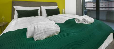 A hotel room with a large bed, green and white bedding, two artworks on the wall, and a view through the window.