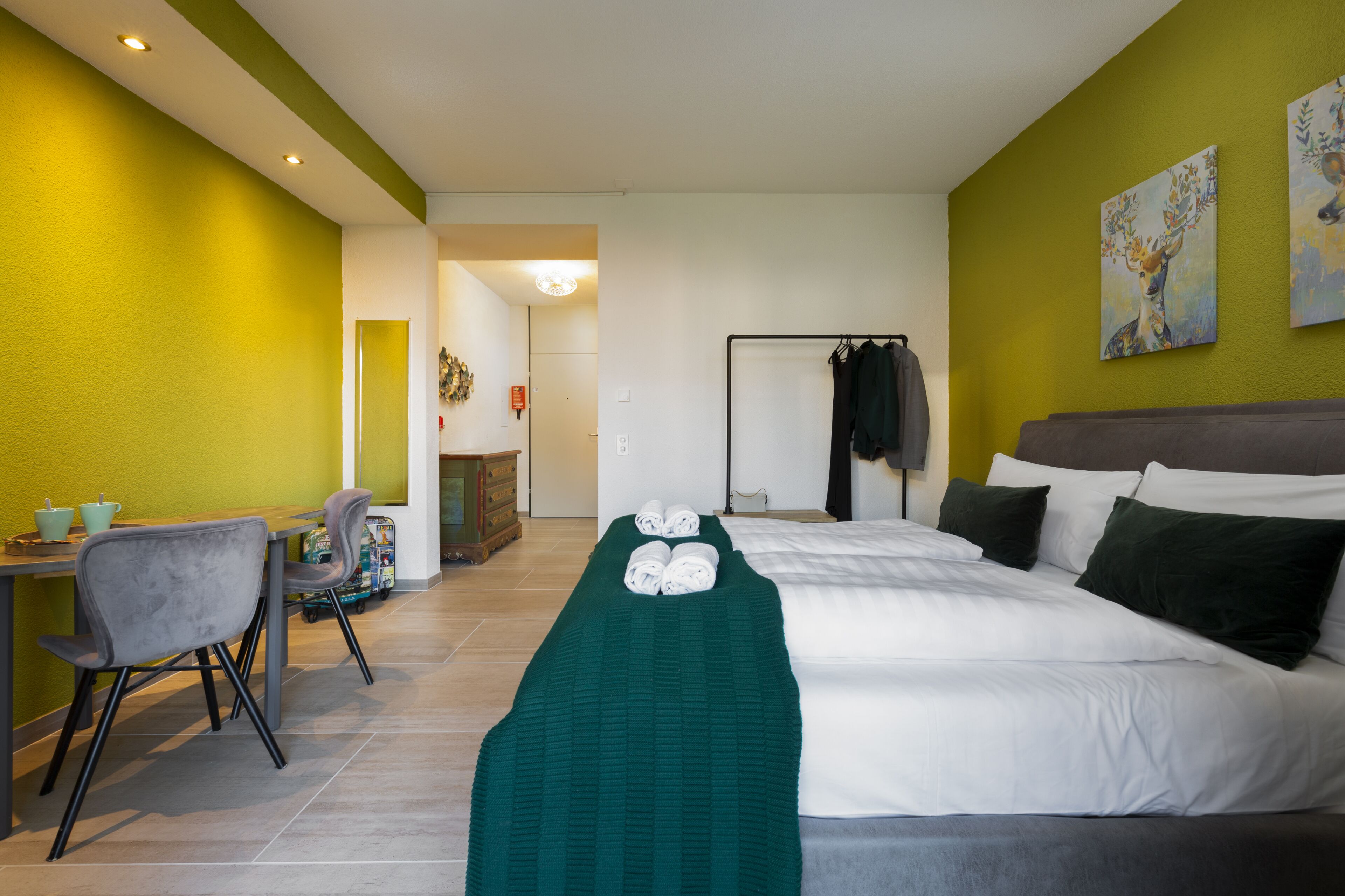 Habitación de hotel con una cama, un escritorio con sillas, un sujetador de toallas verde y obras de arte en las paredes.