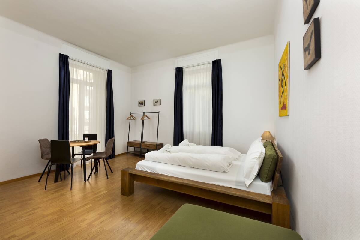 A room with a bed, a desk, chairs, and a hanging coat rack.