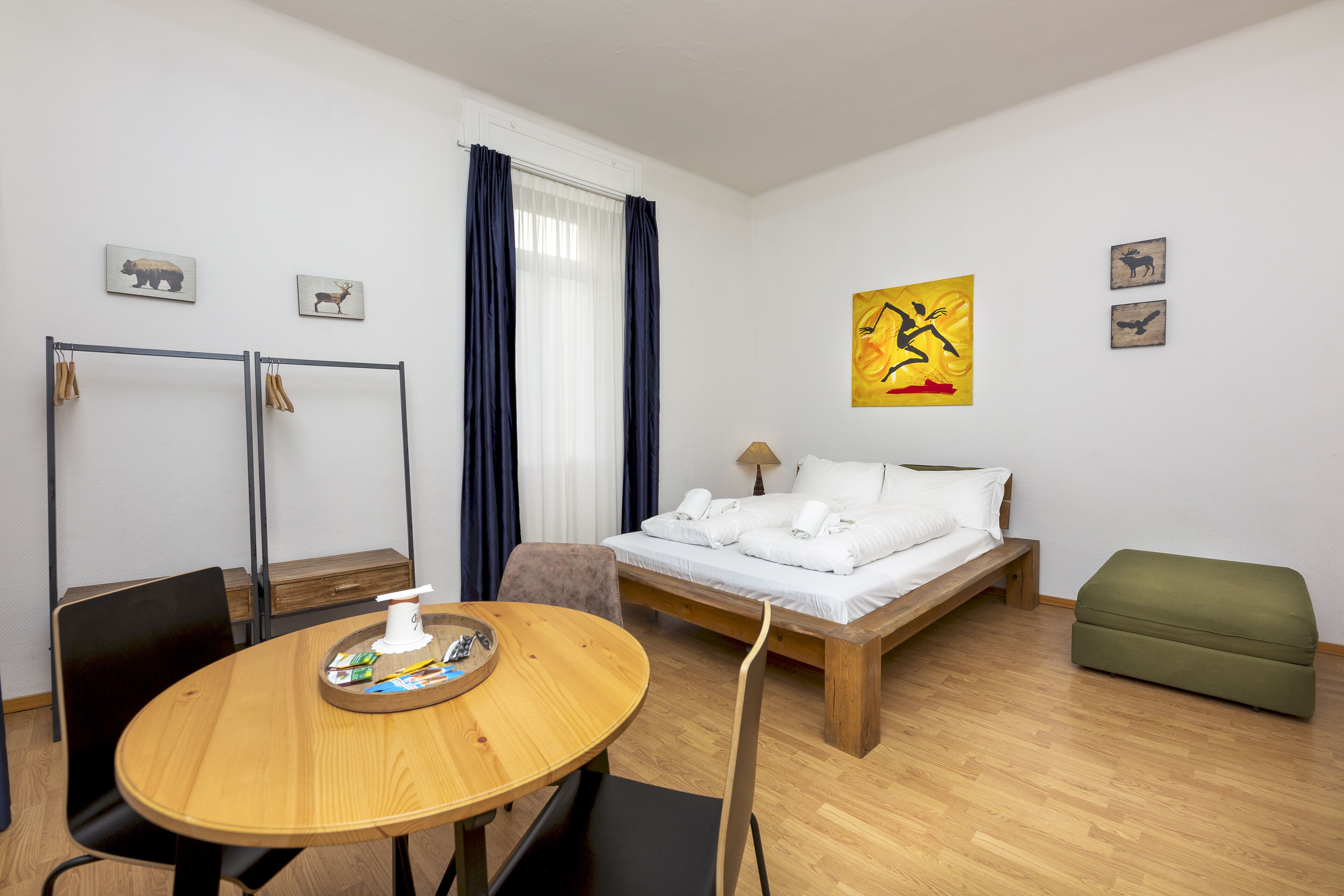 Une chambre d’hôtel avec un lit en bois, un pouf vert, une table à manger en bois avec des chaises, une petite table avec un plateau et une tasse, et une fenêtre avec des rideaux.