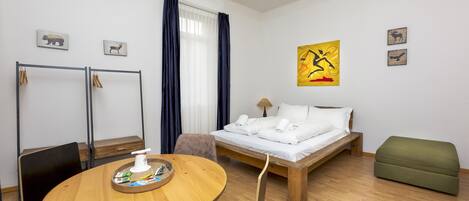 Una habitación de hotel con una cama de madera, un reposapiés verde, una mesa de comedor de madera con sillas, una mesita con una bandeja y una taza, y una ventana con cortinas.