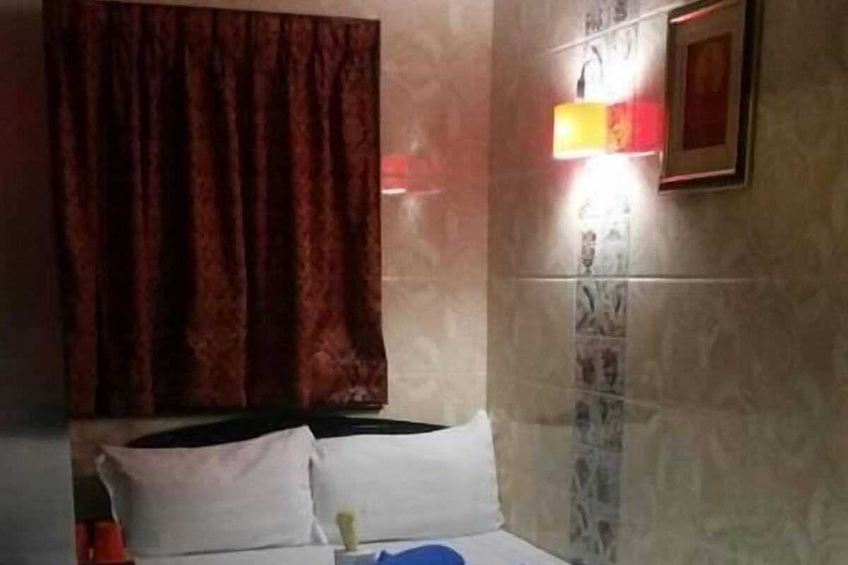 Une chambre d’hôtel avec un lit, une table de chevet, un rideau rouge, une lampe fixée au mur et un sol carrelé.