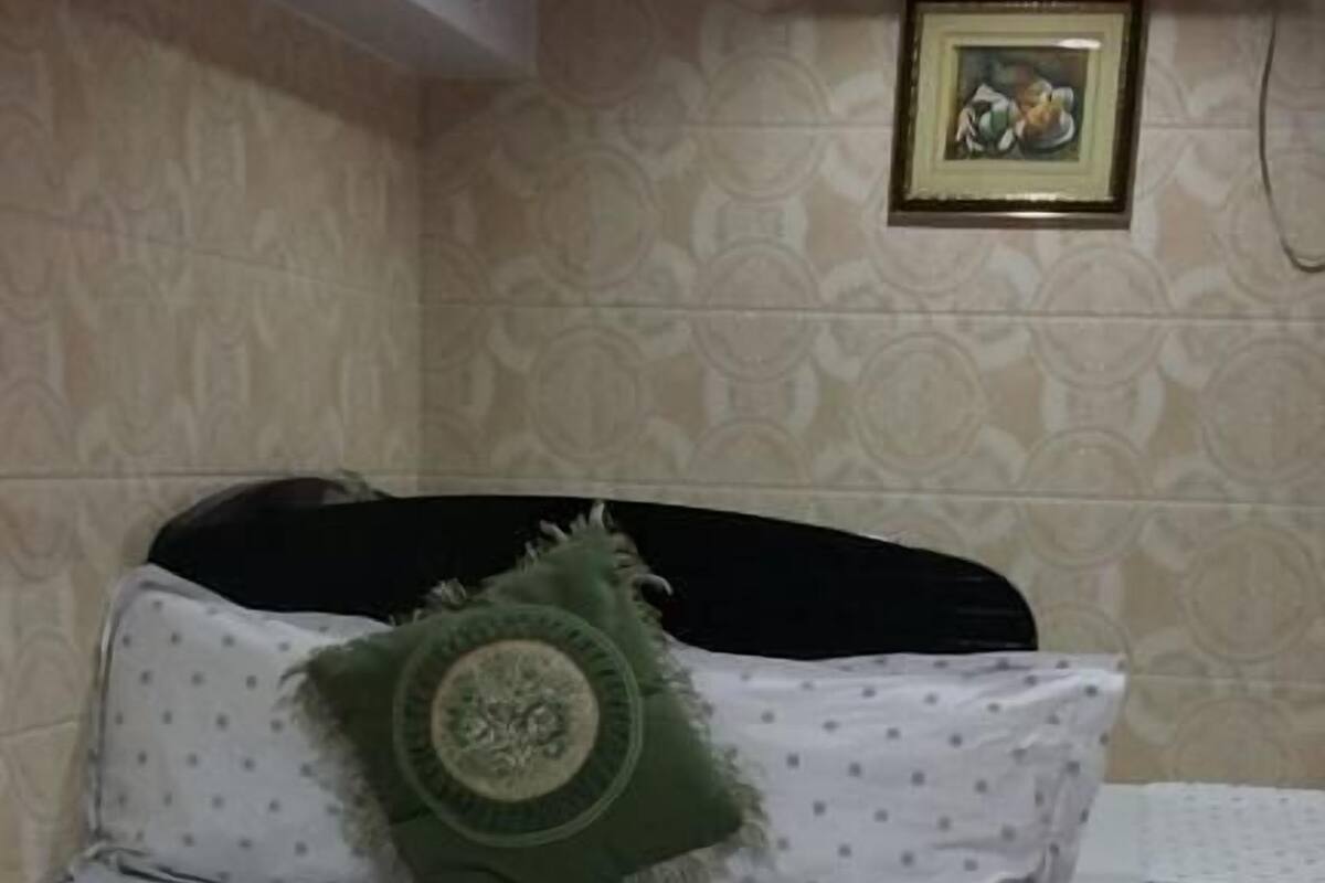 Un lit recouvert d’une courtepointe blanche et orné d’un coussin vert, une petite table recouverte d’une nappe blanche et un climatiseur fixé au mur.