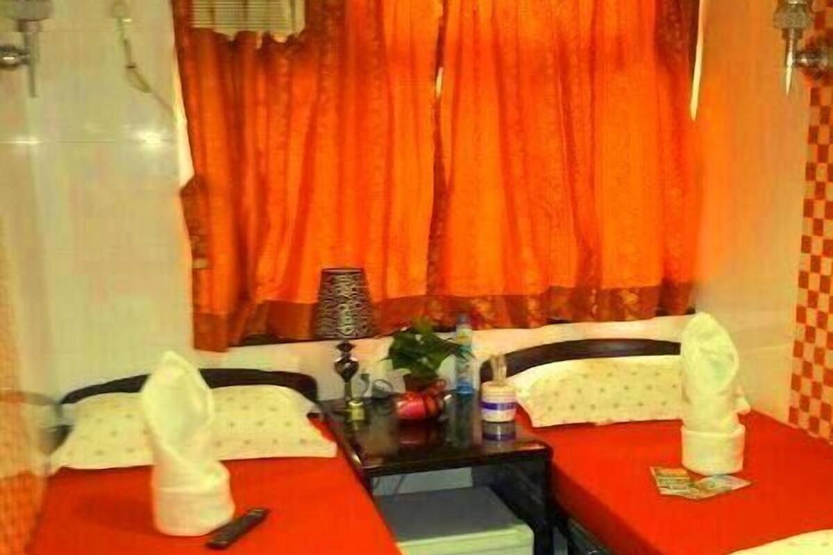 Une chambre d’hôtel avec deux lits simples, une petite table avec une plante, un radiateur et une fenêtre avec des rideaux orange.
