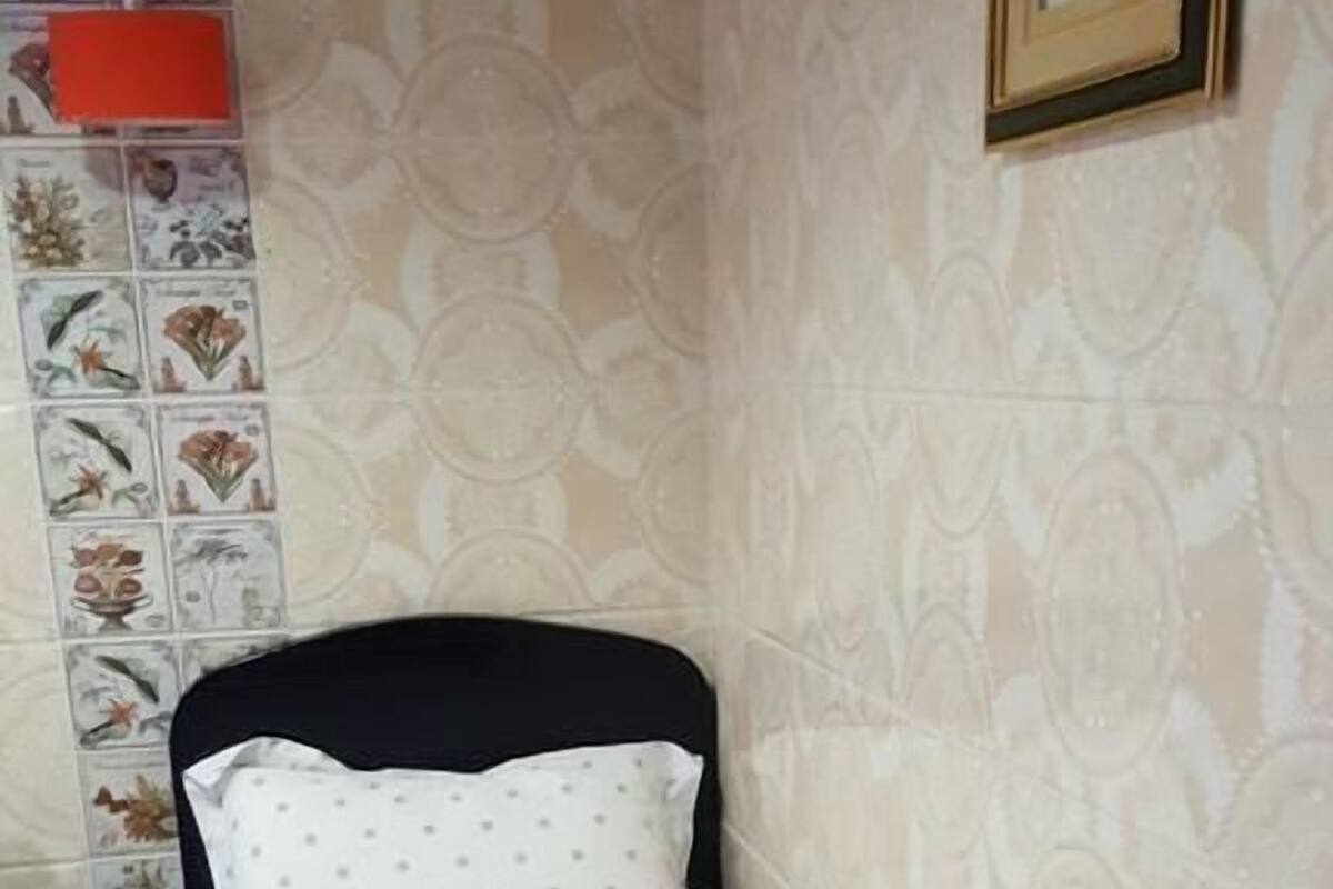 Un petit lit bien fait, avec un drap blanc et un seul oreiller, une tête de lit noire et un mur orné d’un motif floral.